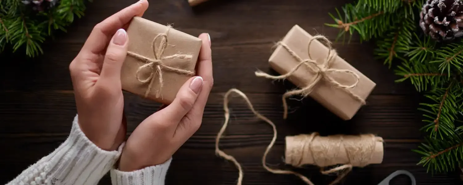 Mãos preparando presentes de Natal sustentáveis com embalagens eco-friendly, fita de juta e decoração com ramos de pinheiro, promovendo inovação em embalagens verdes.
