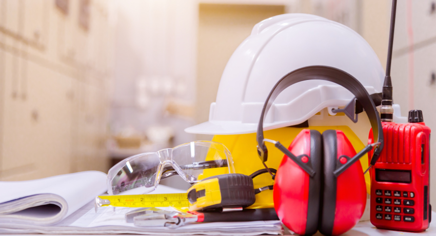 Equipamentos de segurança do trabalho, como capacete, óculos de proteção, protetor auricular e rádio comunicador, sobre mesa com plantas técnicas.