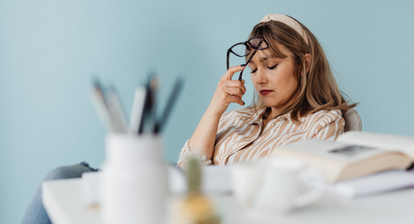 Mulher com blusa listrada sentada à mesa segura os óculos e toca a testa, com expressão de cansaço; há livro e xícara sobre a mesa.