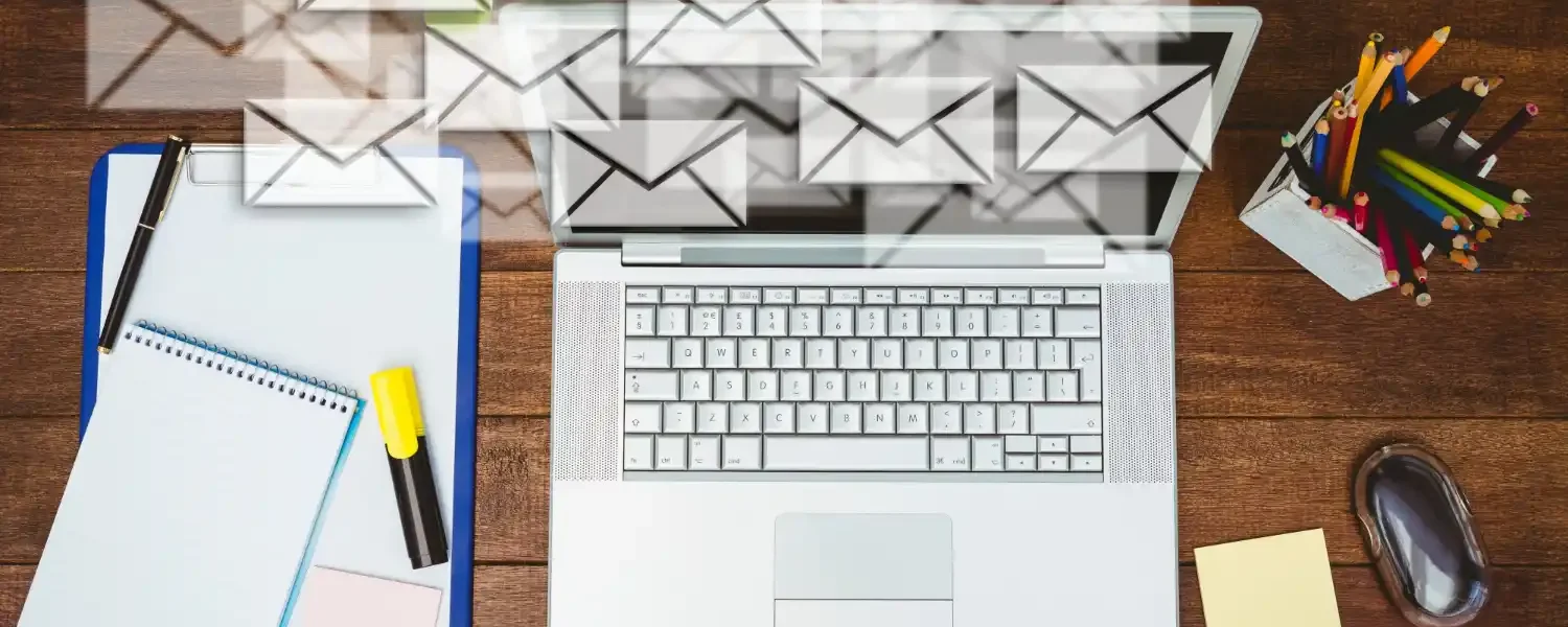 Mesa de trabalho com laptop aberto e ícones de e-mails sobrepostos, além de caderno, caneta e lápis, em cenário que representa E-mail Marketing em 2026: Estratégias para Aumentar Engajamento e Conversões.