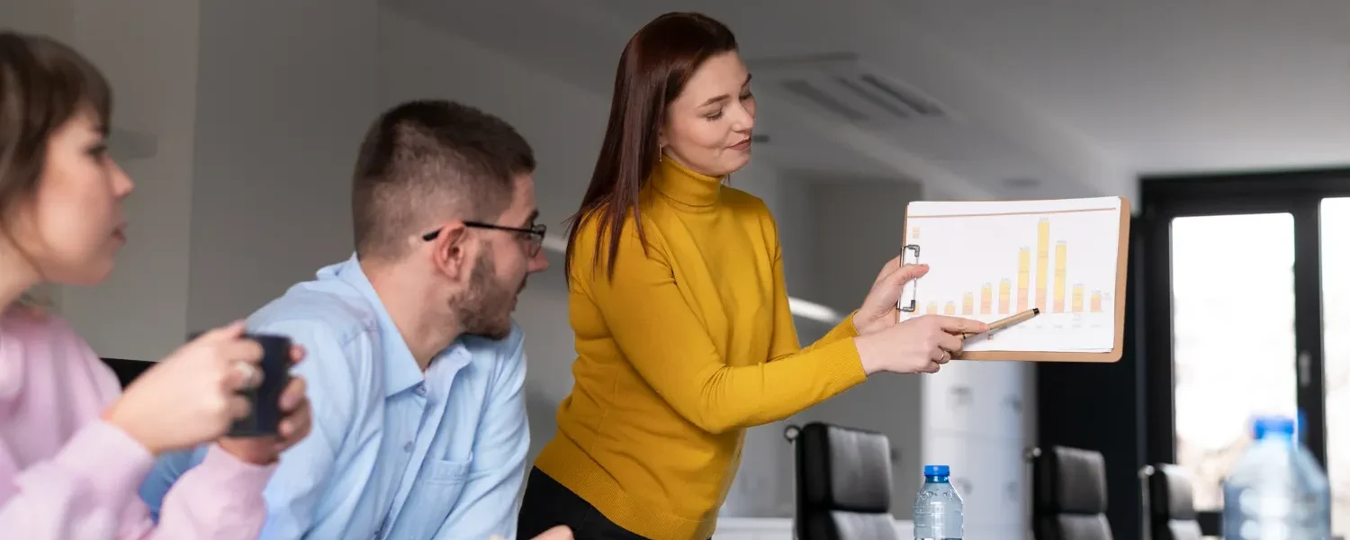 Marketing de Performance: Como Transformar Dados em Decisões Estratégicas em uma reunião, uma mulher mostra um gráfico em um tablet enquanto colegas analisam resultados em uma sala de negócios.